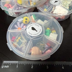 May include: Clear plastic divided containers filled with miniature trinkets. The containers have a circular shape with multiple compartments. The trinkets include small figurines, flowers, and other decorative items. The containers have a label that says "TRINKET TROVE TRADE COLLECT PLAY".