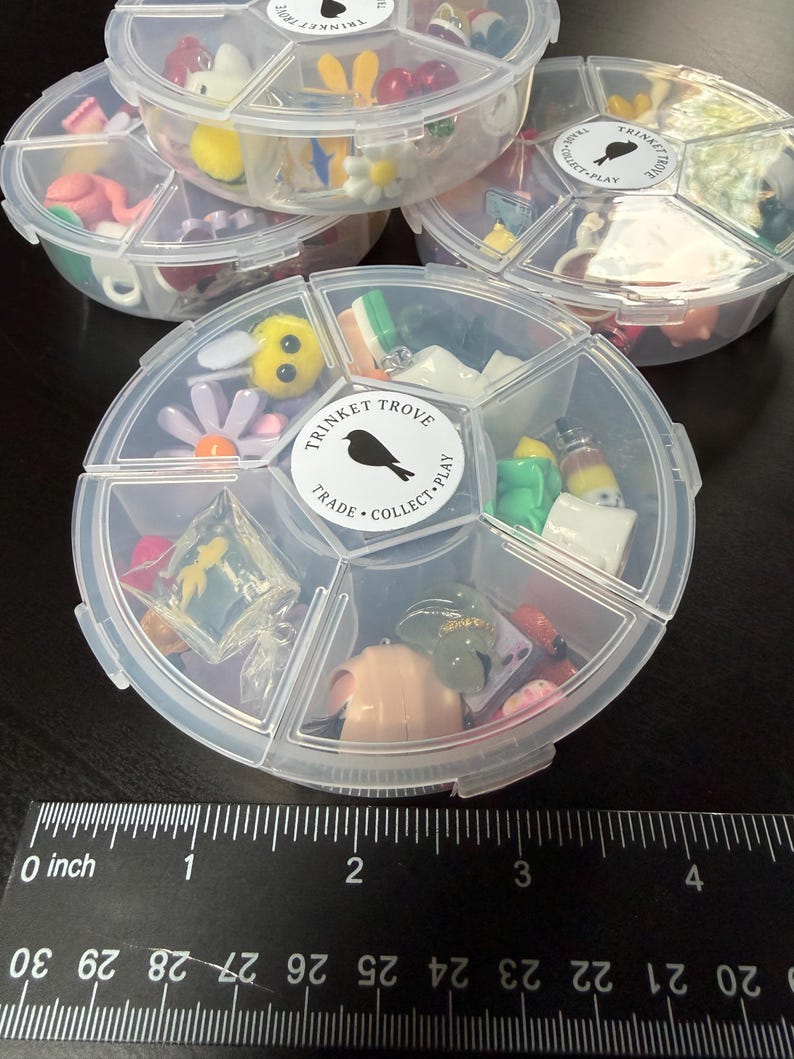 May include: Clear plastic divided containers filled with miniature trinkets. The containers have a circular shape with multiple compartments. The trinkets include small figurines, flowers, and other decorative items. The containers have a label that says "TRINKET TROVE TRADE COLLECT PLAY".