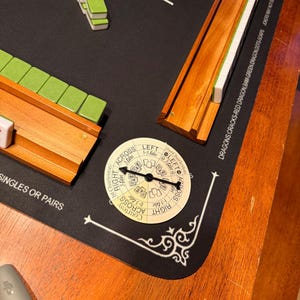 May include: A Mahjong game set with green and white tiles, wooden racks, and a circular game piece. The game is set on a black mat with white text and decorative corner designs. The wooden table is visible.