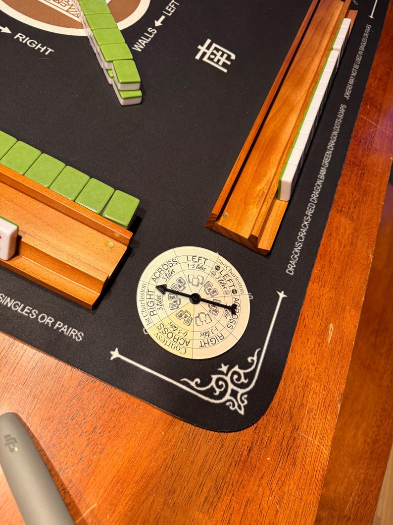 May include: A Mahjong game set with green and white tiles, wooden racks, and a circular game piece. The game is set on a black mat with white text and decorative corner designs. The wooden table is visible.