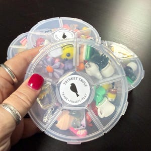 May include: Clear plastic organizer filled with colorful trinkets, including flowers, bees, and miniature objects. The organizer has a round shape with multiple compartments. The label reads "TRINKET TROVE, TRADE, COLLECT, PLAY" with a bird silhouette.