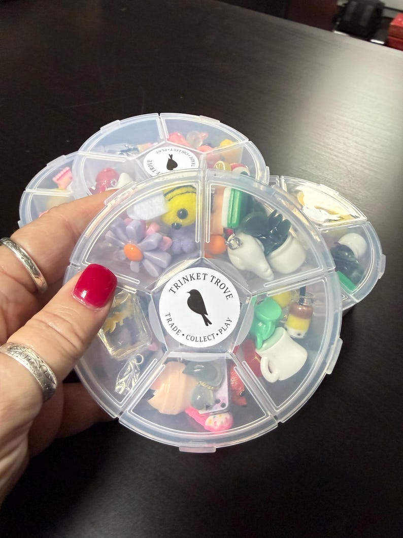 May include: Clear plastic organizer filled with colorful trinkets, including flowers, bees, and miniature objects. The organizer has a round shape with multiple compartments. The label reads "TRINKET TROVE, TRADE, COLLECT, PLAY" with a bird silhouette.