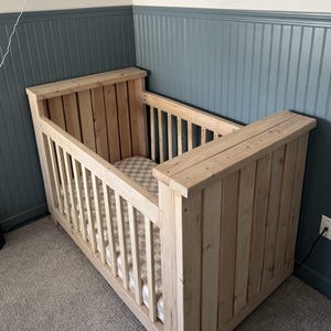 May include: A wooden crib with a slatted design and a white and tan checkered mattress. The crib is made of natural wood and has a simple, rustic style.