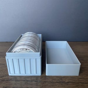 May include: A light blue rectangular container holding a stack of silver coins, alongside an empty container. The coins have text on them. The containers are on a wooden surface.