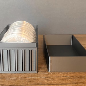 May include: A grey rectangular storage box with a lid, next to an empty box. The box contains a stack of silver coins. The box has a ribbed design on the front. The empty box is a dark grey colour.