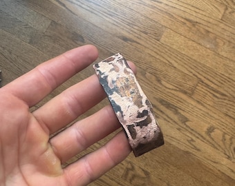 May include: A wide, rigid cuff bracelet made of metal with a textured, mottled surface. The exterior features a mix of dark, oxidized patches and bright, polished copper-toned areas, creating a high-contrast, weathered appearance across the band.