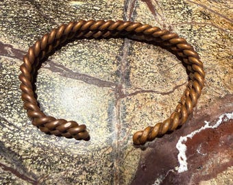 May include: A copper-colored cuff bracelet featuring a twisted rope design. The bracelet is circular with an open gap at the front, showcasing a metallic finish with a textured, braided appearance throughout the entire length of the band.