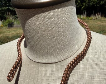 May include: A copper-colored necklace featuring a twisted rope design. The necklace is open-ended with two distinct, spiraled strands that curve around the neck, showcasing a metallic finish and a uniform, textured, braided appearance throughout.