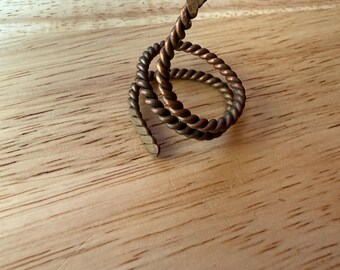 May include: A copper-colored metal ring crafted from a single piece of twisted wire. The ring features a spiral design that wraps around itself, with one end tapering to a point and the other end featuring a flattened, textured finish.