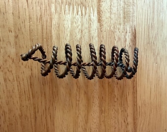 Puede incluir: Un objeto en espiral de metal marrón oscuro construido con alambre trenzado. El diseño presenta un núcleo central de alambre recto envuelto con un alambre trenzado continuo y estrechamente enrollado, creando una serie de bucles uniformes.