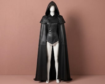 May include: A black leather hooded cape featuring a structured bodice with a pointed hemline and ribbed shoulder detailing. The garment includes a deep hood and a long, flowing cloak that extends to the floor, worn over a white mannequin torso.