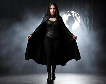 May include: A woman in a black outfit, including a long cape, leather corset, and pants, stands in a dramatic pose. The cape is open, revealing the outfit. The background is dark and smoky, with a spotlight effect.