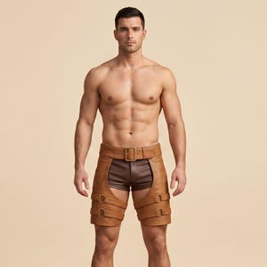 May include: Tan leather chaps featuring a wide waistband with a central buckle and two adjustable buckled straps around each thigh, worn over dark brown shorts. The chaps have a structured design with visible stitching and metal hardware.
