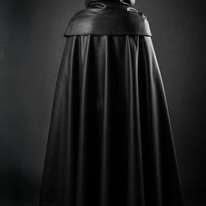 May include: A black hooded cloak, viewed from the back, with a smooth, leather-like texture. The cloak has a wide, flowing design, and the hood is up. The image is set against a dark background, emphasizing the garment's silhouette.