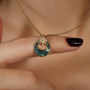 May include: Gold-toned necklace featuring a pendant with a crescent-shaped, iridescent green and blue inlay. A starburst design with clear crystals sits above the crescent. The necklace is displayed on a finger.