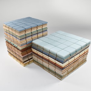 May include: Two stacks of square tiles in various colours. The top layer of each stack is a light blue, with a grid pattern. The sides of the stacks reveal a mix of colours, including beige, brown, and green. The tiles appear to be made of ceramic or a similar material.