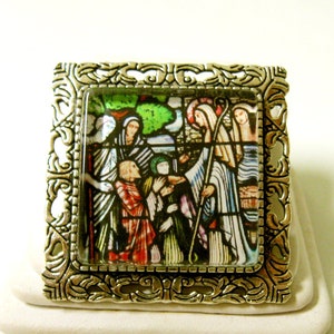 May include: A silver-toned metal frame with an ornate design surrounds a colourful stained glass image of people in a religious scene.
