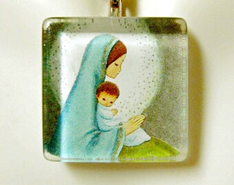 Puede incluir: Un collar de cadena de plata con un colgante cuadrado de vidrio que presenta una pintura de María sosteniendo al niño Jesús. La pintura está en una suave gama de colores azul, verde y blanco.