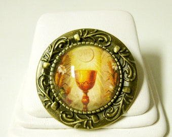Op de afbeelding: Een gouden metalen pin met een sierlijk ontwerp. Het midden van de pin heeft een glazen koepel met een religieus beeld van een kelk en een hostie.