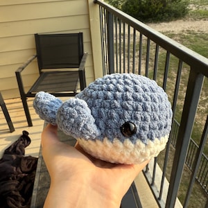 May include: A handmade crochet whale plushie in shades of blue and white. The whale has a textured, soft appearance with a black eye. The plush toy is a cute, handcrafted item.