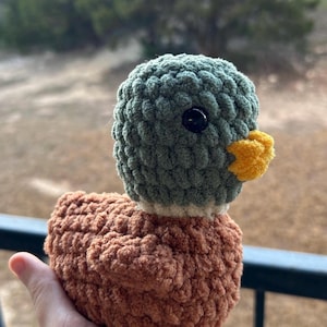 May include: A crocheted stuffed duck toy with a green head, brown body, and yellow beak. The duck has black eyes and a white stripe around its neck.