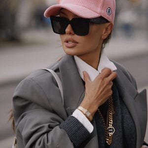 Może przedstawiać: Kobieta w różowej sztruksowej czapce bejsbolowej, czarnych okularach przeciwsłonecznych i szarej marynarce. Ma również na sobie złoty zegarek i złoty łańcuszek. Tło jest rozmyte.