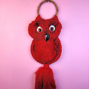 May include: A handmade red owl wall hanging with a bamboo handle. The owl has large, round eyes with white and black details, a black beak, and a fluffy tassel. The owl is made of a textured, possibly felted material.