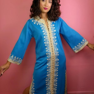 May include: A bright blue kaftan featuring intricate gold embroidery along the neckline, front placket, and sleeve cuffs. The garment includes a vertical row of small gold buttons down the center front and side slits at the hemline.