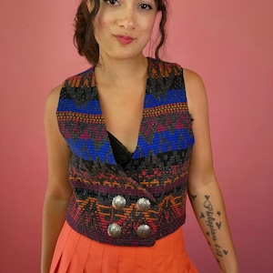 May include: A sleeveless vest featuring a multi-colored geometric pattern in shades of blue, orange, and grey. The vest has a V-neckline, a double-breasted front closure, and four round, silver-toned metal buttons with an embossed floral design.