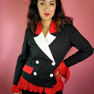 May include: A black blazer with a white lapel and red accents. The blazer features a double-breasted design with white buttons and red ruffled trim at the cuffs and hem. The background is a solid pink.