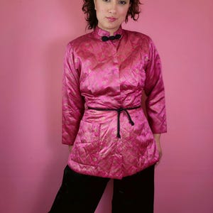 Puede incluir: Chaqueta acolchada rosa con cuello mandarín y mangas largas. La chaqueta tiene un estampado dorado y está ceñida a la cintura con un cinturón de cuerda negro. La persona lleva pantalones negros.