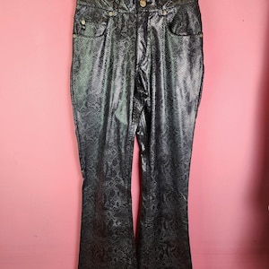 May include: Black, flared trousers with a snakeskin pattern. The trousers are hanging on a black hanger against a pink background. They have a high waist, pockets, and a button closure.
