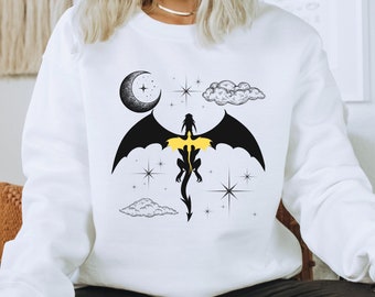Puede incluir: Sudadera blanca con una silueta de dragón negro volando sobre un fondo de luna creciente, nubes y estrellas. Las alas del dragón están extendidas y su cola está enroscada. El cuerpo del dragón está delineado en amarillo.