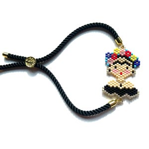 Könnte beinhalten: Ein Armband aus schwarzem Kordel mit einem goldenen Verschluss und einem Perlenanhänger von Frida Kahlo mit einem bunten Blumenkranz.