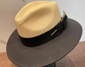 May include: A tan and brown fedora hat with a black band and a small feather accent. The hat is sitting on a black and silver stand.