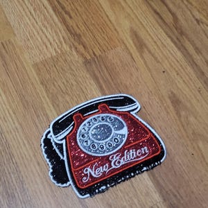Könnte beinhalten: Ein roter, schwarzer und weißer Aufnäher in Form eines alten Wählscheibentelefons. Das Telefon hat eine glitzernde rote Oberfläche und eine schwarze Umrandung. Der Text "New Edition" ist auf dem Telefon geschrieben.