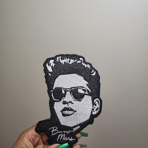 May include: Black and white embroidered patch featuring a stylized portrait of a person wearing sunglasses. The patch has the text "Bruno Mars" in cursive. The patch is black with white embroidery.