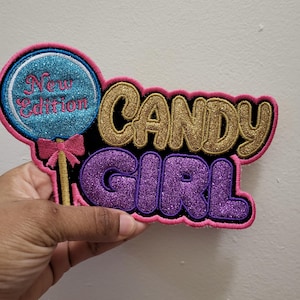 Puede incluir: Un parche colorido con las palabras "CANDY GIRL" en letras doradas y moradas brillantes. El parche también presenta una piruleta azul con las palabras "New Edition" y un lazo rosa.