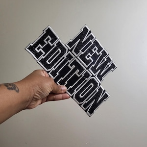 May include: Black and white embroidered patches spelling out "NEW EDITION" in a bold, collegiate font. The patches are held in a hand against a neutral background. The patches have a glittery texture.