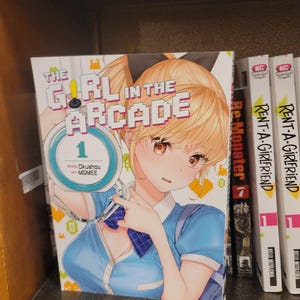 Op de afbeelding: Een manga-volume getiteld "The Girl in the Arcade" met een blonde anime-stijl karakter op de cover. De cover heeft gepixelde tekst en een blauw-wit kleurenschema. Andere manga-volumes zijn zichtbaar op de plank.