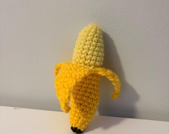 May include: A handmade crochet banana with a yellow peel and a yellow and tan body. The banana peel is partially open, revealing the banana inside. The banana has black tips.