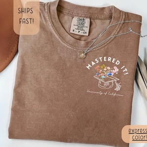May include: Brown t-shirt with the text "MASTERED IT!" and a graduation cap design with flowers. The shirt also has the text "University of California" and the year "2025". The shirt is a "expresso color!"