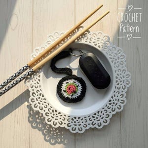 May include: A crocheted sushi keychain with a black, white, and pink design, next to a black car key fob and bamboo chopsticks with floral patterned ends, all on a white decorative plate. The plate has a scalloped edge. The words "CROCHET Pattern" are in the top right corner.