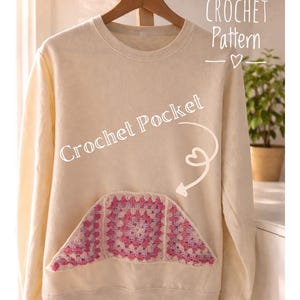 May include: A cream-coloured sweatshirt featuring a crocheted pocket in pink and white. The pocket is a granny square design. Text on the sweatshirt reads "Crochet Pocket" and "Crochet Pattern". It hangs on a wooden hanger.
