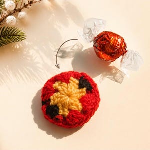 May include: A round, crocheted cover in red, yellow, and black yarn, positioned below a red-wrapped Lindt Lindor chocolate truffle. A thin, dark brown curved arrow points from the chocolate toward the circular crocheted item.