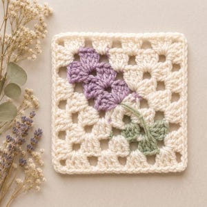 Puede incluir: Un posavasos cuadrado de ganchillo tipo granny square con una base de hilo color blanco roto y un motivo floral central. La flor está tejida con hilo morado claro para los pétalos e hilo verde salvia para el tallo y las hojas integradas.