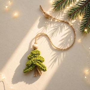 May include: A green knitted Christmas tree ornament featuring a textured yarn structure, a small wooden bead at the top, and a natural jute twine loop for hanging. The tree has a small brown jute tassel at the base, creating a simple, handcrafted look.