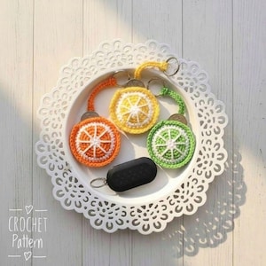 May include: A collection of handmade crochet keychains shaped like citrus fruit slices in orange, yellow, and green. Each keychain has a silver ring and a matching colored strap. A black car key fob rests on a white, decorative tray.