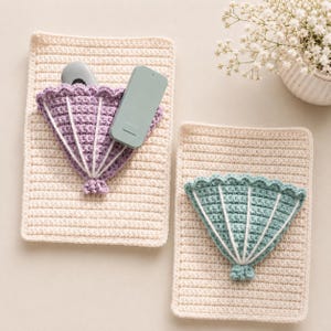 May include: Two rectangular cream-coloured crochet wall organisers, each featuring a fan-shaped pocket. One pocket is lavender and the other is sage green, both accented with white vertical stitching and scalloped edges. A remote is inside the lavender pocket.