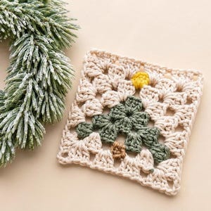 May include: A square crochet coaster featuring a cream-colored background with a central dark green tree motif, a small yellow star at the top, and a brown trunk at the base, crafted with a traditional granny square stitch pattern.